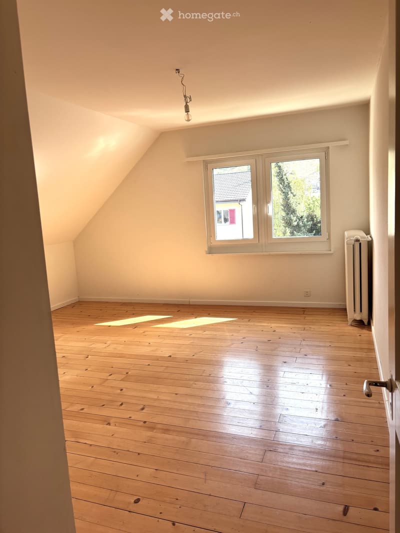 Empty attic room with wooden flooring, white walls, and a single window offering a view of the outside. A radiator is visible on the right side.
