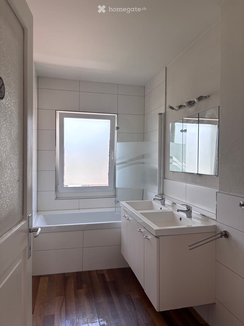 Modern bathroom with a bathtub, double sink, large mirror, and a window providing natural light.