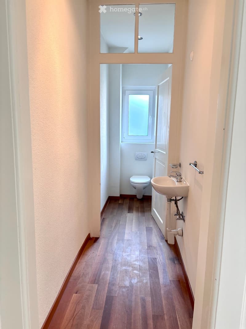 A narrow hallway with wooden flooring and light-colored walls, leading to a small bathroom with a toilet and sink.