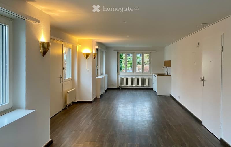 Empty hallway with hardwood floors, white walls, windows, and wall lamps. A small kitchenette with a sink is at the far end of the hallway.