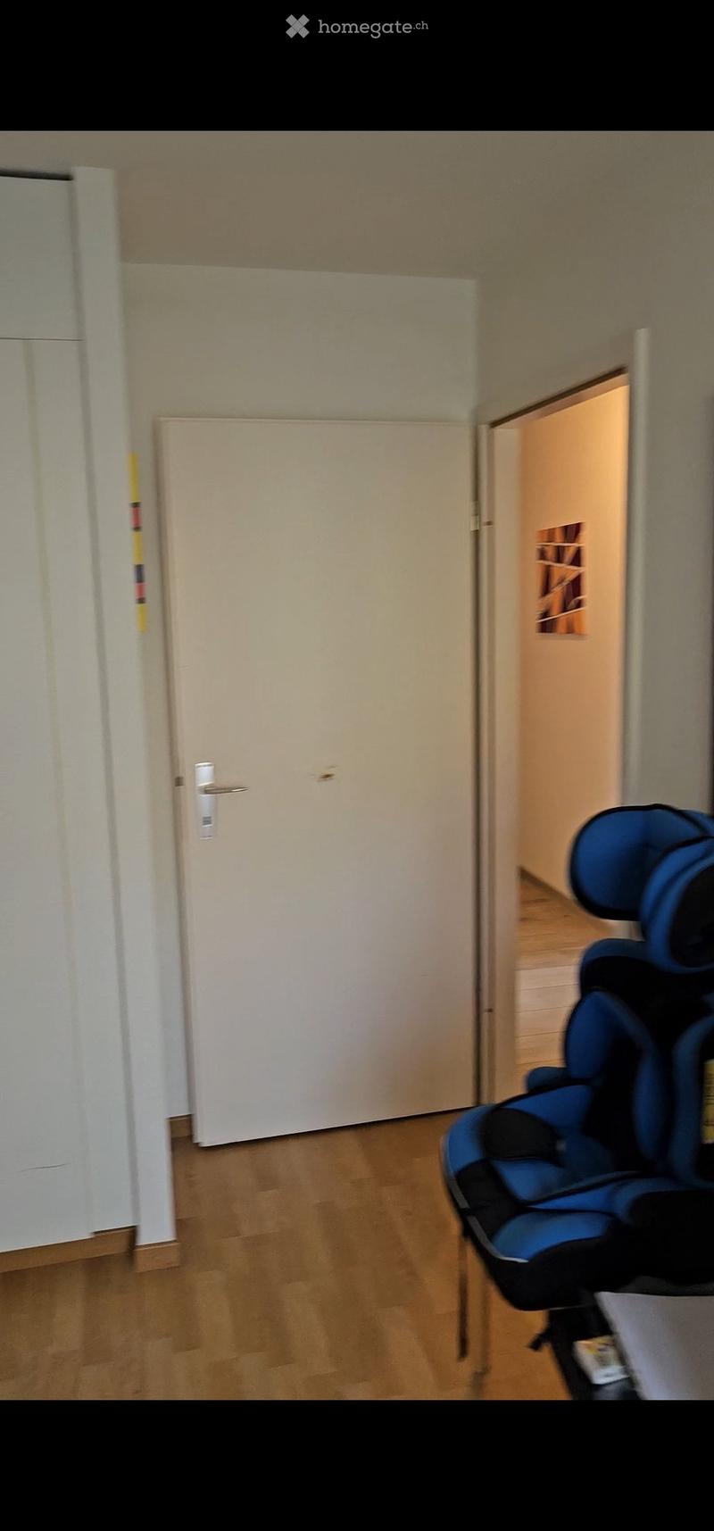 A small, unfurnished room with wooden flooring and a closed white door. A blue baby chair is positioned near the door.