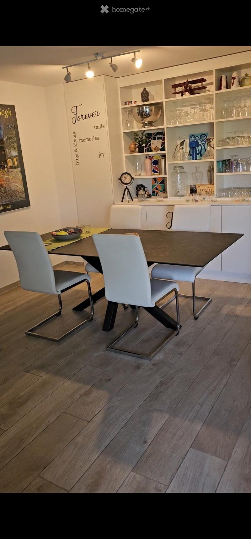 Modern kitchen with a dining area, featuring a dark wooden dining table, white chairs, and built-in cabinets with glass doors displaying decorative items and glassware.