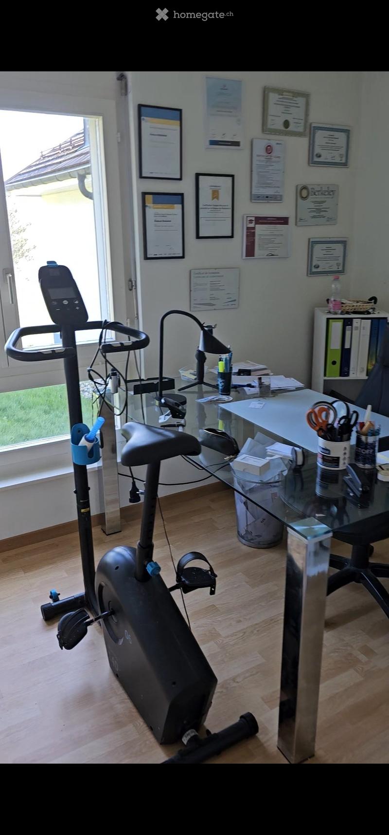 A well-lit room with wooden flooring, featuring an exercise bike, a glass desk with office supplies, a chair, a lamp, and several framed certificates on the wall. A large window offers a view of greenery outside.