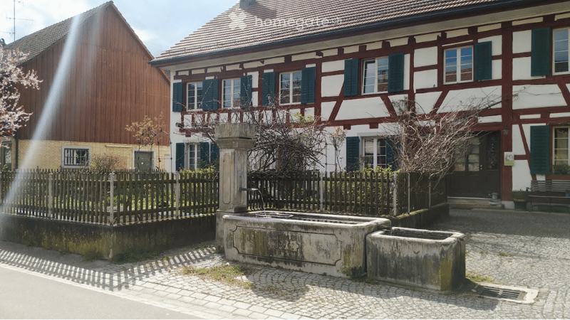 Traditional half-timbered house with white and red wooden framing, multiple windows with shutters, and a tiled roof. A stone well and wooden fence are visible in the foreground.