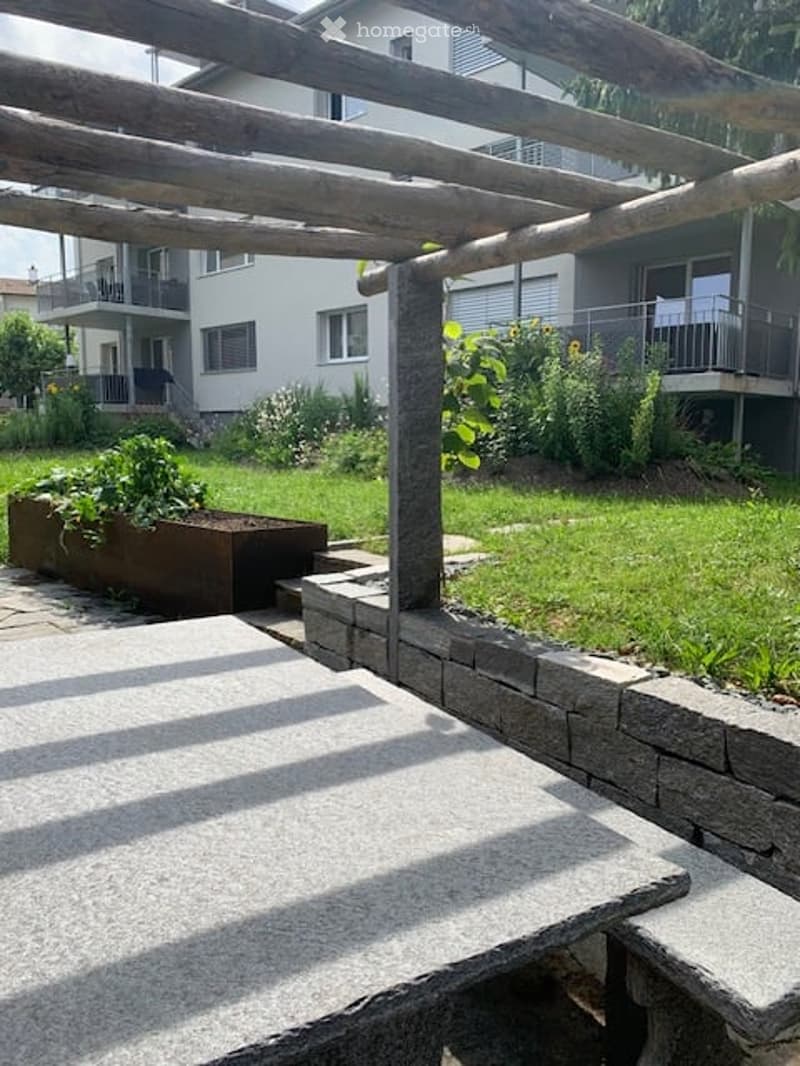 Garden area with a wooden pergola, stone seating, raised planter, and lush greenery.