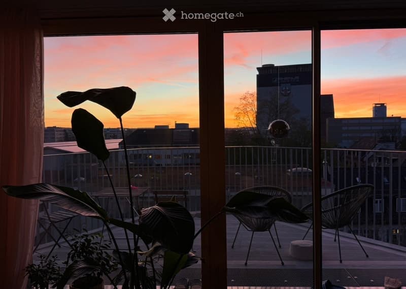 Balcony with wooden decking, metal railing, and cityscape view at sunset. Features include a small table, chairs, a round stool, and indoor plants near the window.
