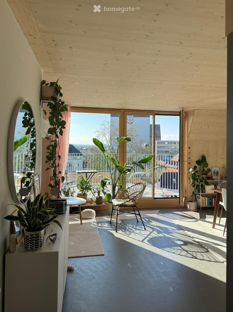 Bright, wooden-floored room with large windows offering a city view, featuring a wicker chair, small table, mirror, bookshelf, and several potted plants.