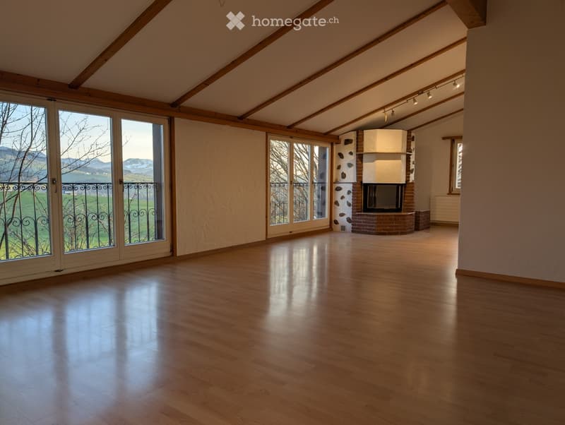 Empty room with wooden flooring, exposed wooden beams, large windows with black wrought-iron railings, and a brick fireplace.