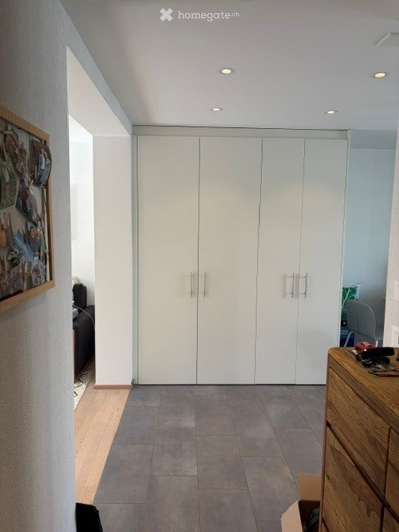A modern, minimalistic room with a large white wardrobe, a wooden drawer unit, and a picture frame on the wall. The floor is tiled, and the walls are painted white.