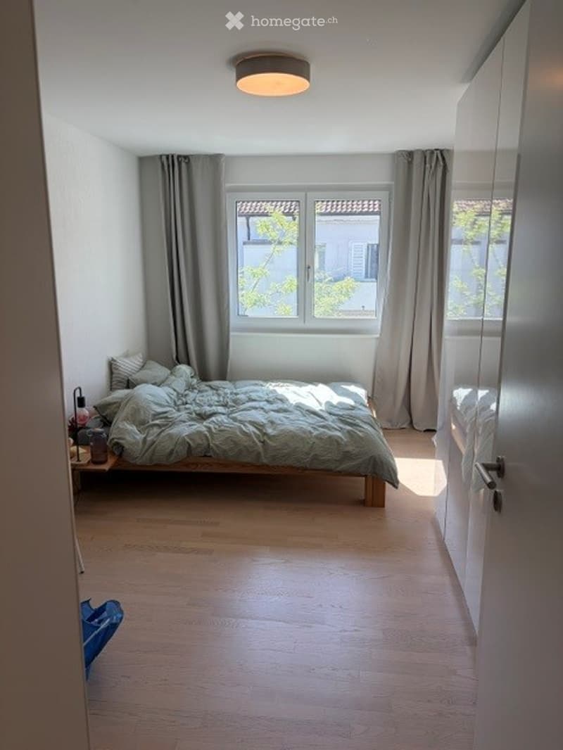 Bright bedroom with wooden flooring, large window with grey curtains, neatly made bed with green duvet, nightstand with lamp, and a small side table.