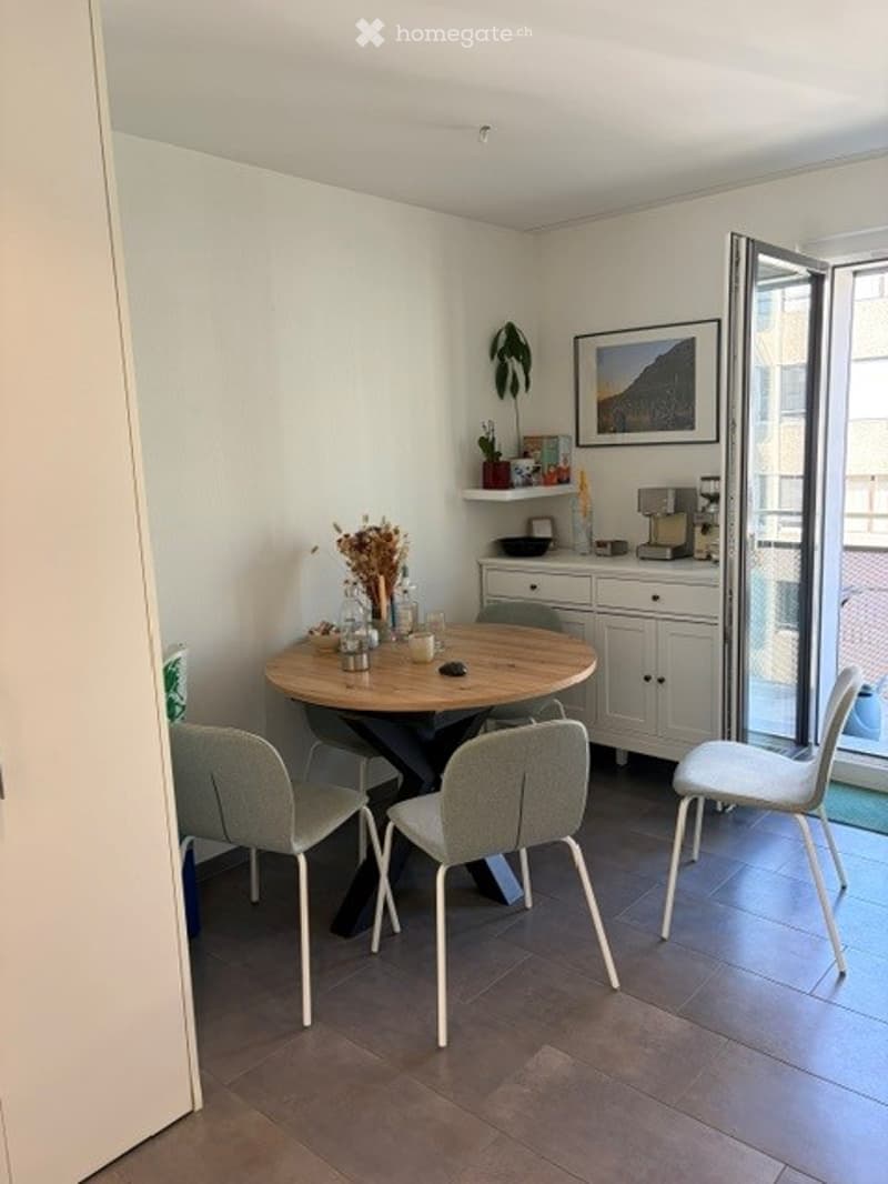 A modern, minimalist dining area with a round wooden table, four grey chairs, a white cabinet, and a coffee machine on the counter. The room features a large window with a view, a small plant, and a picture frame on the wall.