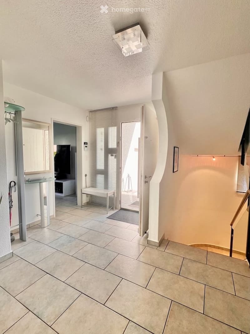 Bright hallway with tiled flooring, white walls, and a modern ceiling light fixture. Features an open doorway leading to another room, a small bench, and a glass console table with a mirror.