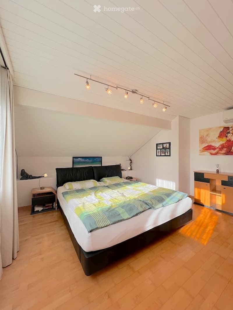 Bright bedroom with wooden flooring, large bed with colorful bedding, nightstand with lamp, desk with chair, and artwork on walls.