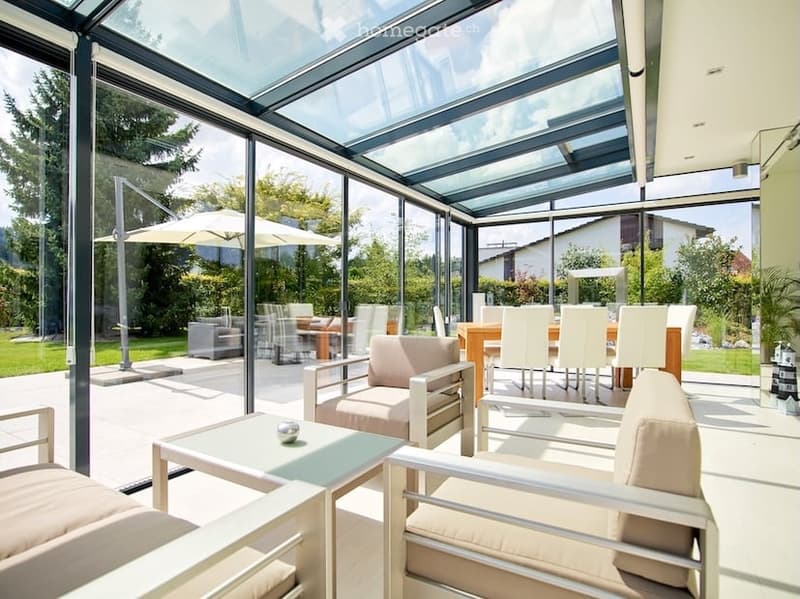 Bright, modern indoor-outdoor living space with large glass walls and a retractable roof, featuring a sofa, chairs, a coffee table, and outdoor dining area with umbrellas.