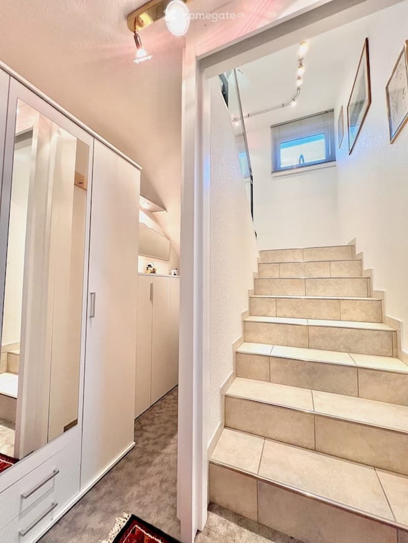 A modern hallway with a staircase leading upwards, white built-in wardrobes, and a small window at the top of the stairs.