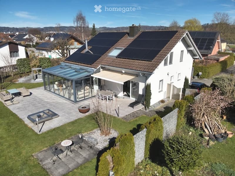 Modern two-story house with solar panels on the roof, surrounded by a well-maintained garden and outdoor seating areas.