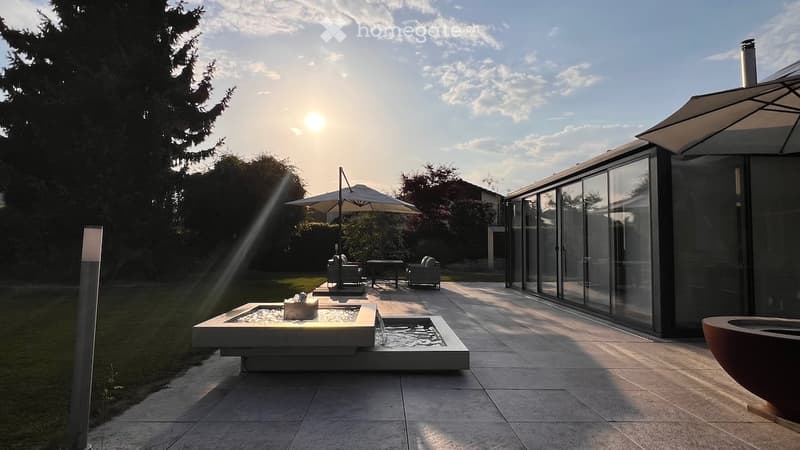 Modern outdoor patio with a water fountain, lounge chairs, umbrella, and sleek glass-walled building.