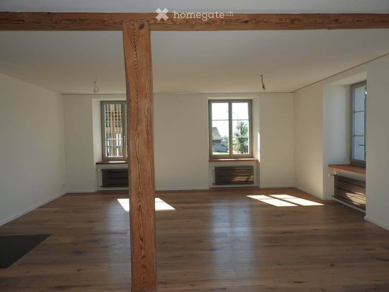 Empty room with wooden flooring, white walls, and large windows allowing natural light. Features a central wooden pillar and radiators beneath the windows.