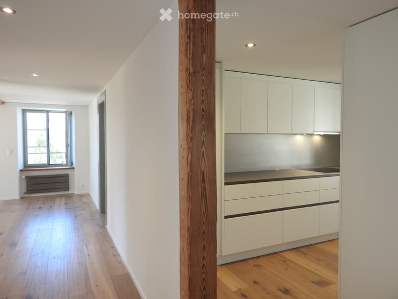 Empty room with wooden flooring, white walls, and a modern kitchen area with white cabinets and stainless steel appliances visible in the background.