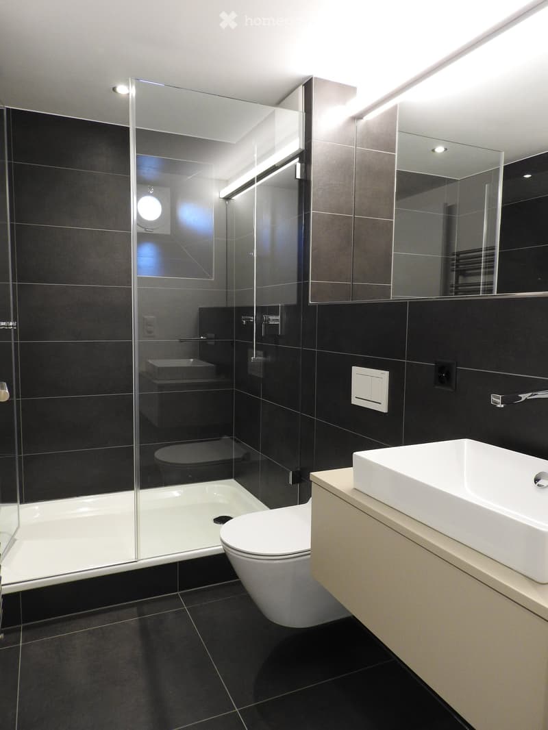 Modern bathroom with black tiles, white bathtub, toilet, and sink, featuring a glass shower enclosure and built-in storage.