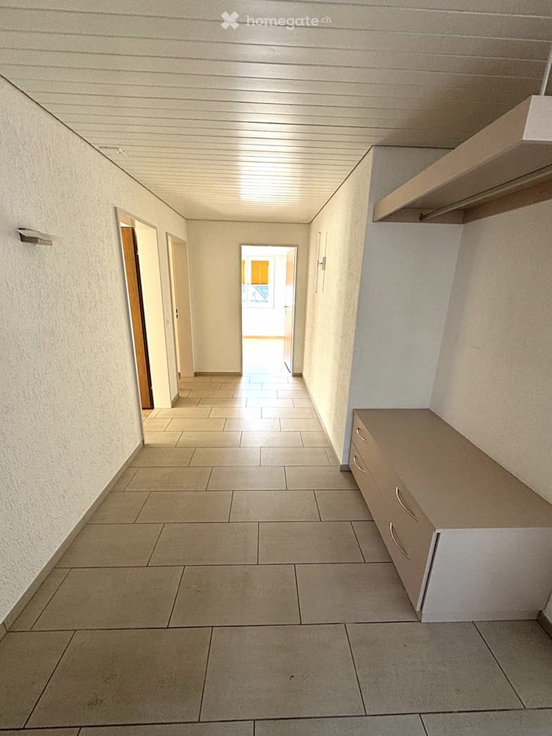 Empty hallway with tiled floor, white walls, and built-in storage unit on the right side. Natural light enters from an open doorway at the end.