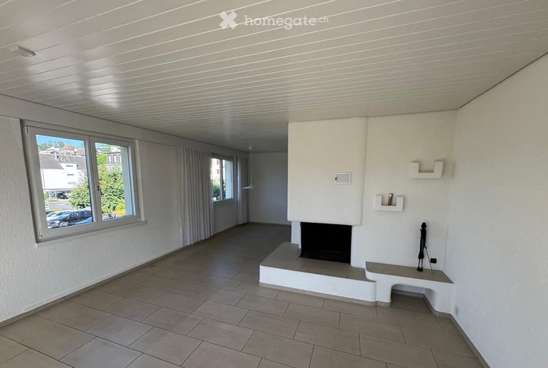 Empty room with tiled floor, white walls, large window with view, fireplace, built-in shelving, and a small bench.
