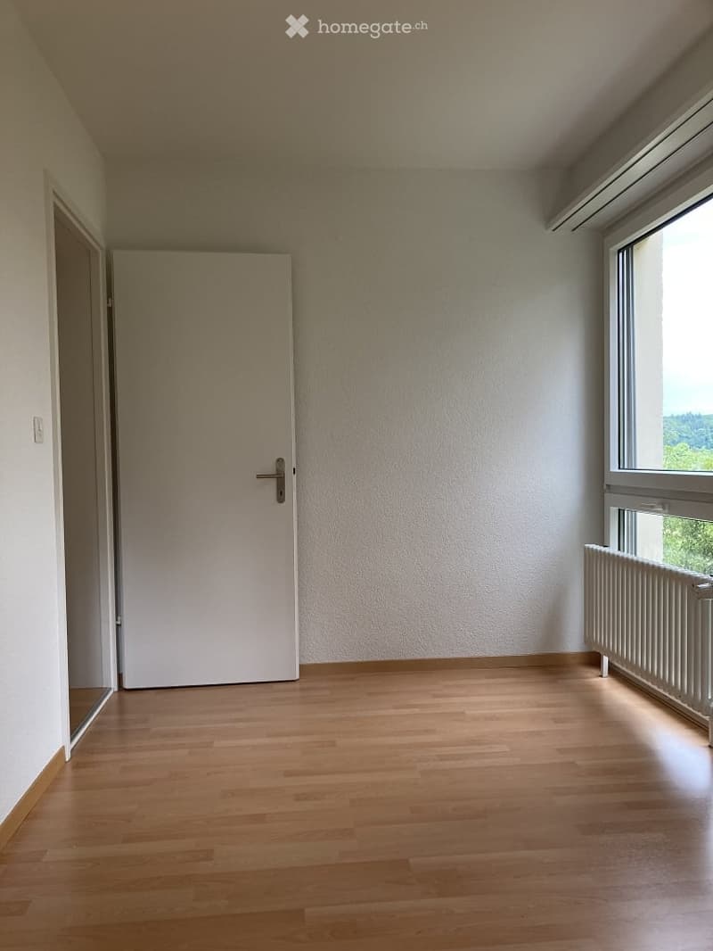 Empty room with wooden flooring, white walls, a door, and a window with a radiator below it.