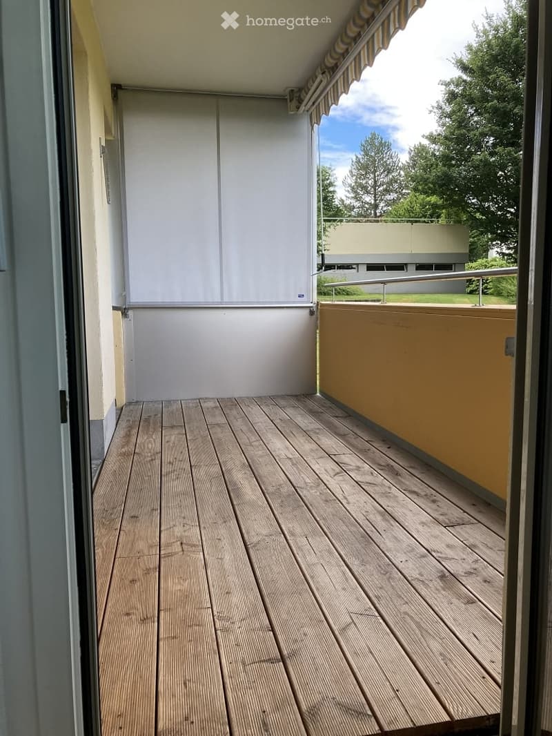Wooden deck balcony with white sliding doors, striped awning, metal railing, and view of greenery.