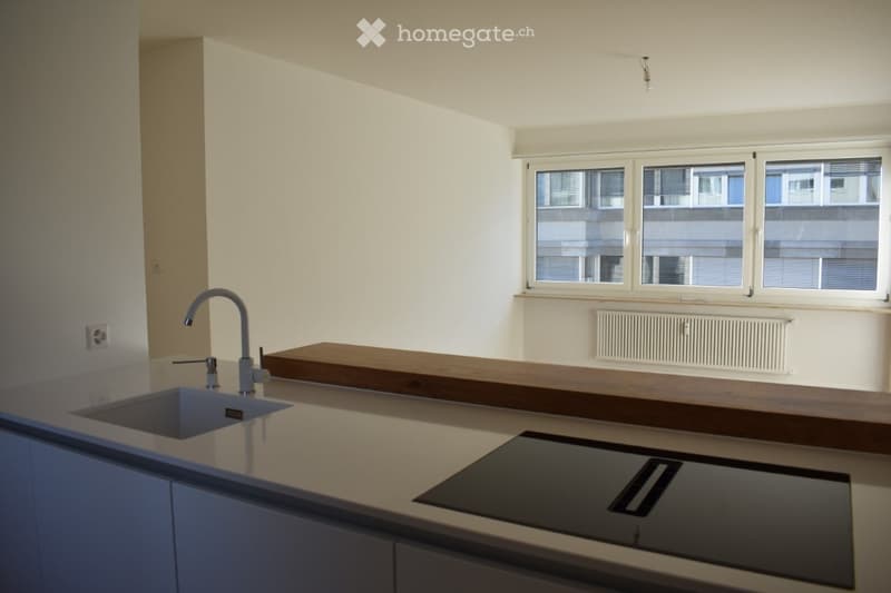 Empty kitchen with modern design, featuring a sink, countertop, and built-in stove. Large windows provide natural light.