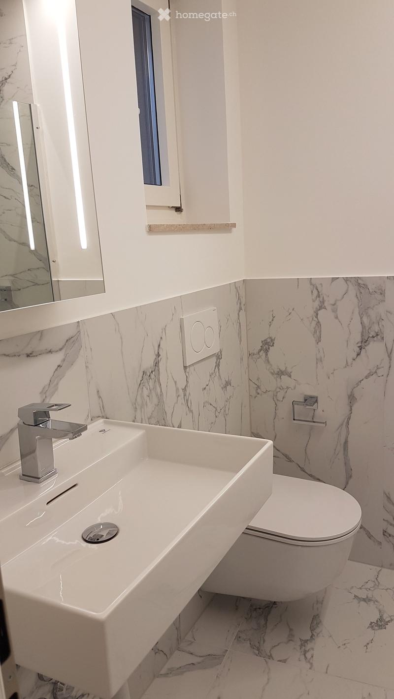 Modern bathroom with white marble tiles, a long sink with a sleek faucet, a toilet, and a large mirror.