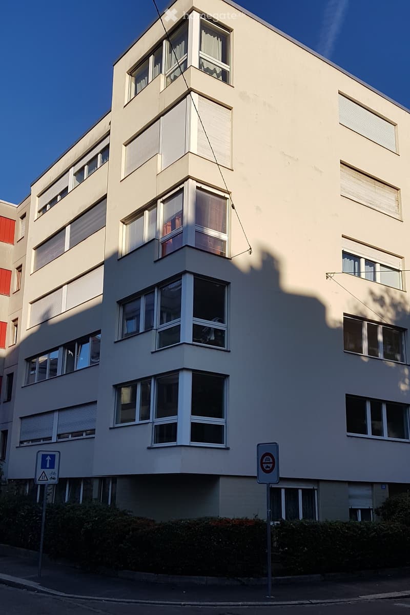 Four-story residential building with light-colored facade, multiple windows with white frames, and some blinds partially drawn. The building has a modern design with clean lines and is situated on a street with traffic signs visible.