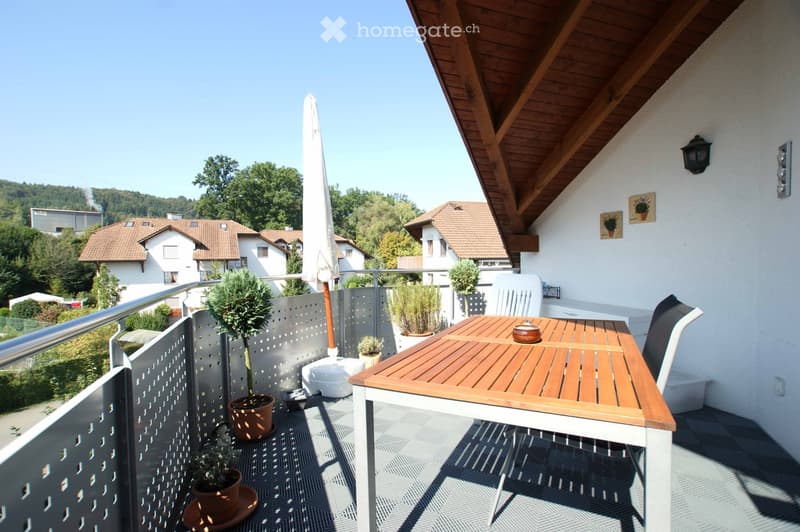 Balcony with wooden table, chairs, potted plants, and an umbrella, overlooking a residential area with houses and greenery.