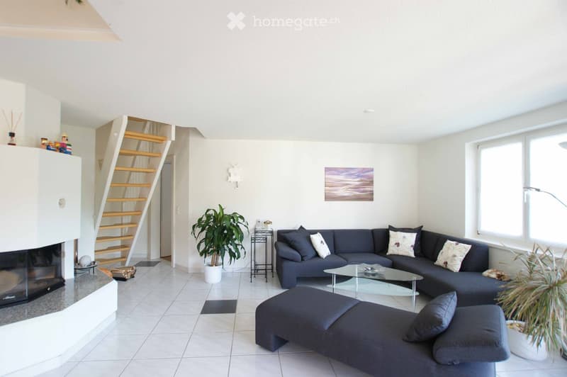 Bright living room with white walls, tiled floor, grey sectional sofa, ottoman, glass coffee table, side table, potted plants, and a fireplace with decorative items on the mantel.