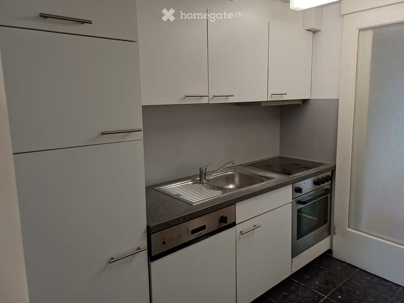 Modern kitchen with white cabinets, stainless steel sink, oven, and refrigerator. Features a dark countertop and tiled floor.