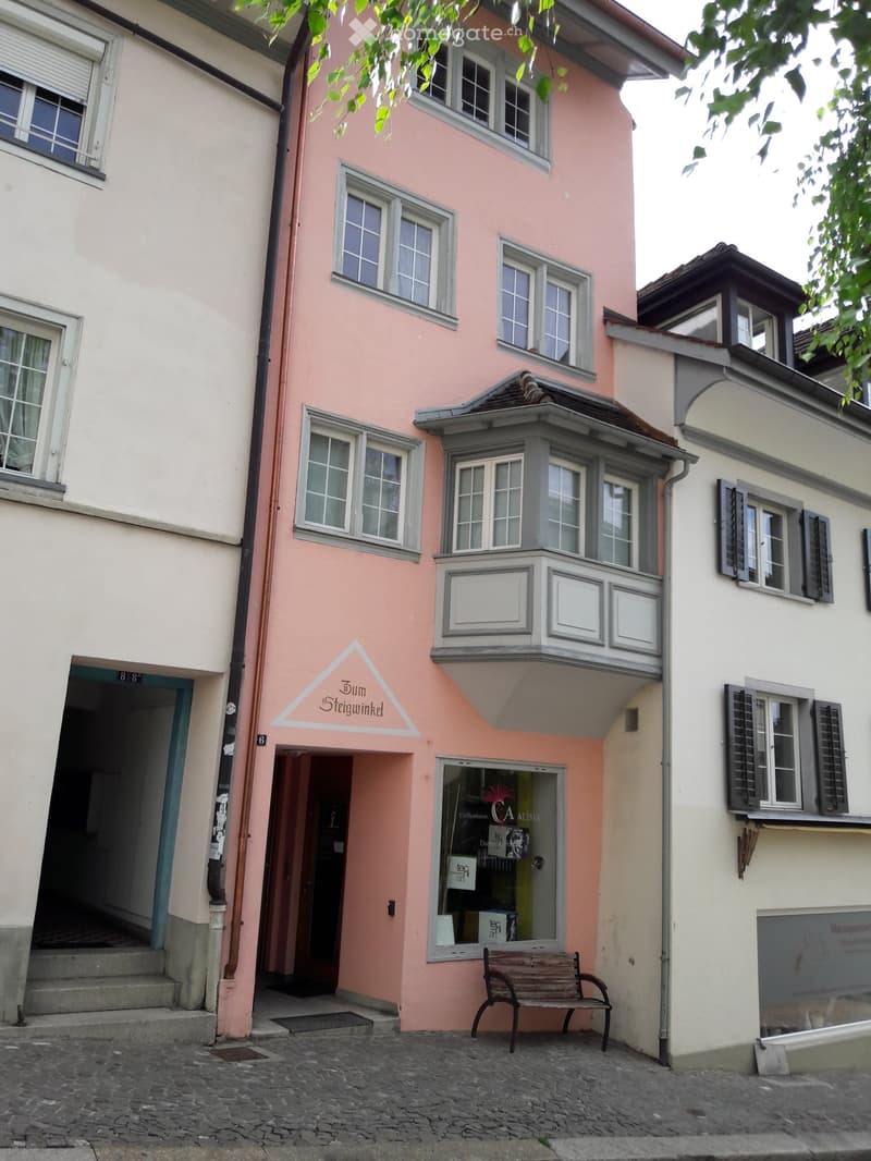A three-story pink building with multiple windows, a balcony, and a storefront with a bench in front.