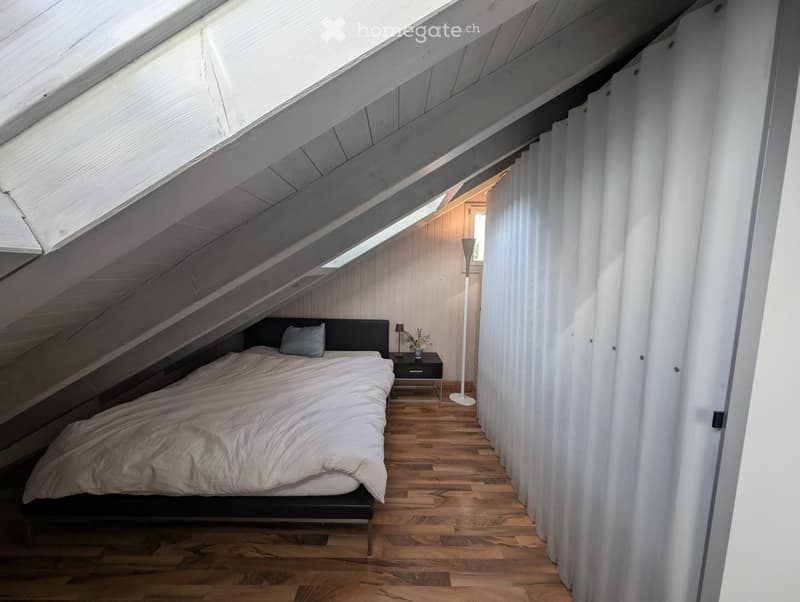 Attic bedroom with wooden floors, slanted ceilings, and skylights. Features a bed with white bedding, a nightstand with a lamp, a floor lamp, and a vase with flowers.
