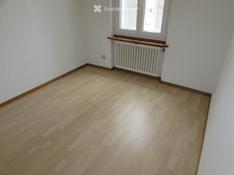 Empty room with light wooden flooring, white walls, and a window with a radiator below it.