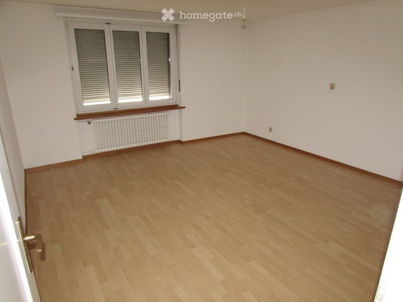 Empty room with light wooden flooring, white walls, and a window with blinds. There is a radiator below the window and a door visible on the left side.
