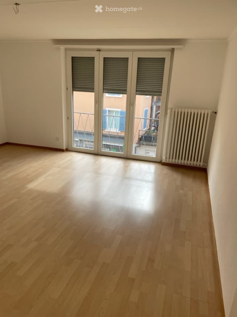 Empty room with wooden flooring, large sliding glass doors leading to a balcony, and a radiator on the wall.