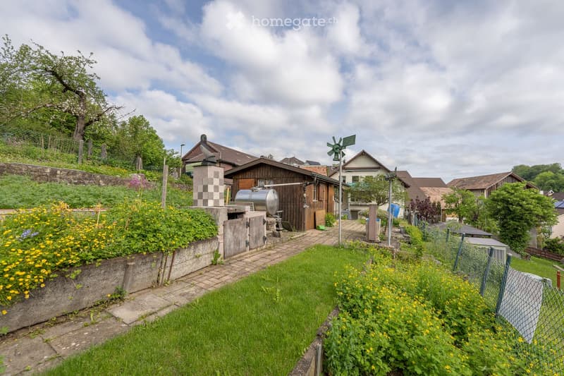 A well-maintained garden with a wooden shed, a water tank, a windmill, and a variety of flowers and greenery. The garden is bordered by a stone wall and a fence, with a paved pathway leading to the shed.