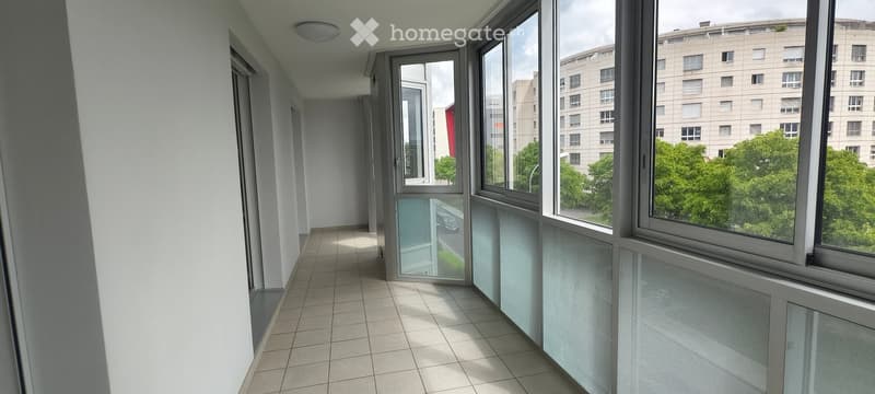 A modern, well-lit hallway with tiled flooring and large windows offering a view of the city and greenery outside.