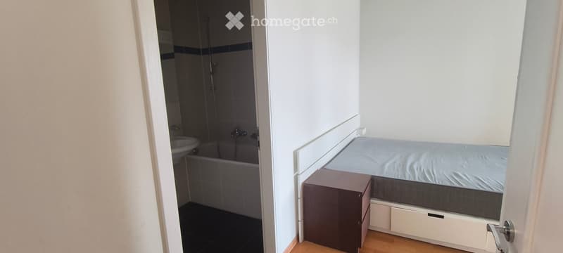 A compact bedroom with a single bed, nightstand, and an adjacent bathroom featuring a bathtub and sink.