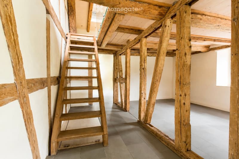 Empty room with exposed wooden beams and a wooden staircase leading to an upper level. The space has light-colored walls and a tiled floor.