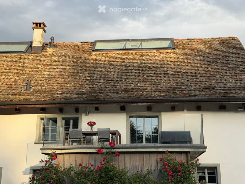 Balcony with a wooden table, two chairs, and a vase of red flowers, attached to a house with a stone roof and multiple windows.