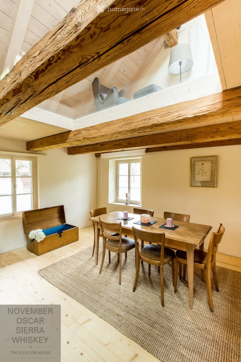 Bright dining area with wooden dining table and chairs, wooden chest with flowers, floor lamp, and candle on windowsill.