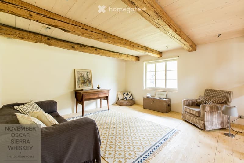Cozy living room with exposed wooden beams, a sofa, armchair, small table, and a lamp. The room features a large window and a patterned rug on the wooden floor.
