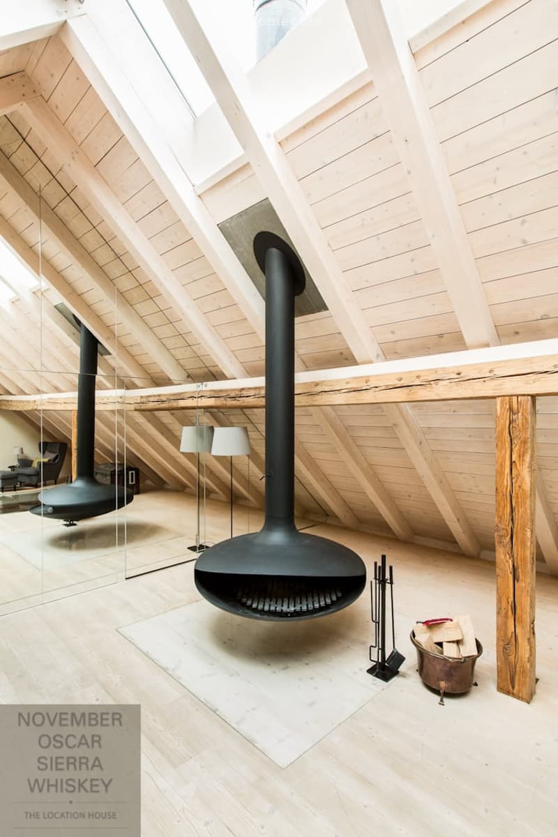 Modern, high-ceilinged room with exposed wooden beams and a large, black, suspended fireplace. The space features a wooden floor, a couple of floor lamps, a small seating area with a chair and table, and a wooden beam as a structural element.