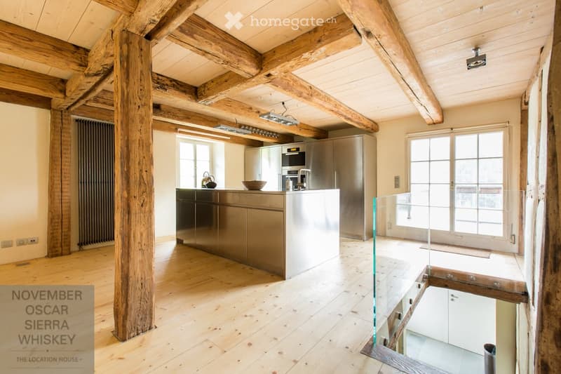 Modern kitchen with exposed wooden beams, stainless steel appliances, and a central island. Large windows provide natural light.