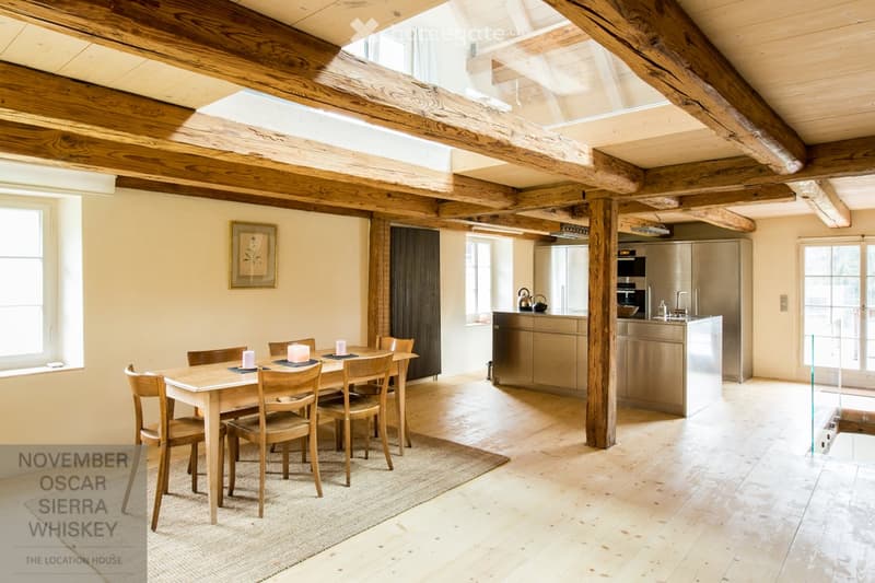 Open-plan living and dining area with exposed wooden beams, a dining table with six chairs, a modern kitchen with stainless steel cabinets, and a framed picture on the wall.