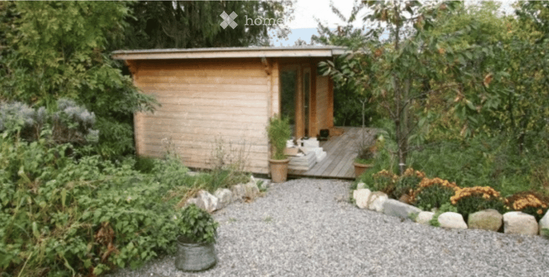 A small wooden cabin surrounded by lush greenery, with a gravel pathway leading to a wooden deck and potted plants.